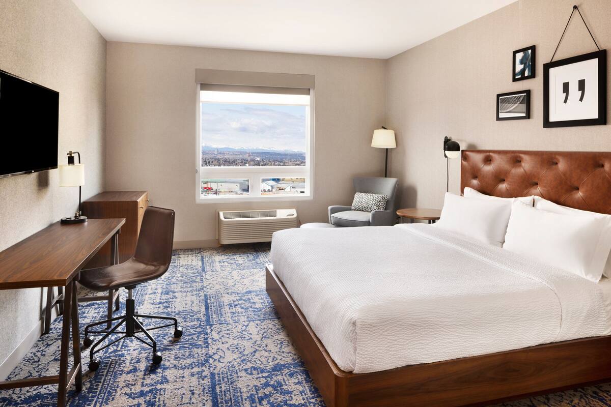 A hotel room with a large bed, a desk, a chair, a TV, and a window with a city view.