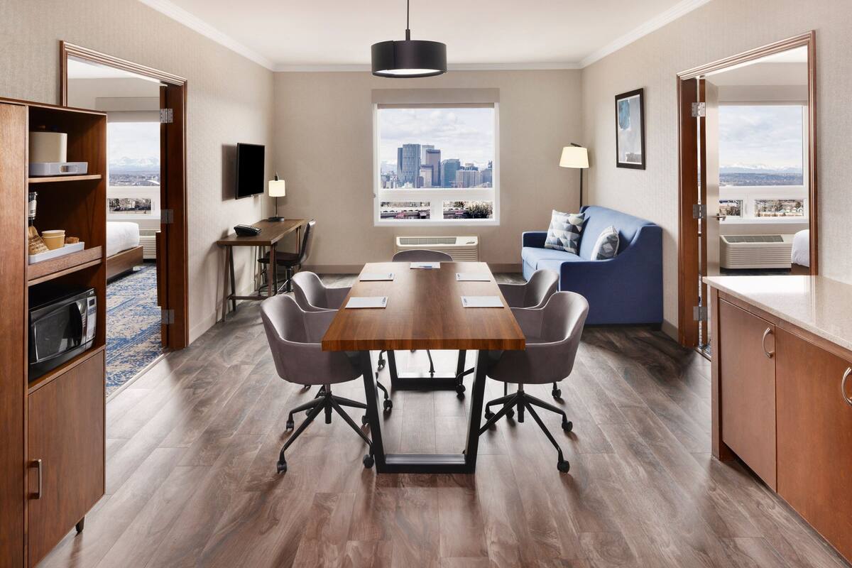 A modern hotel room with a dining area, a sofa, a TV, and a city view.
