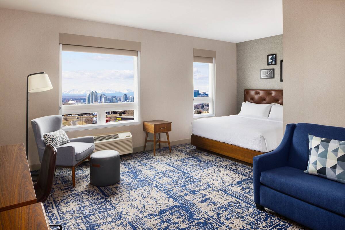 A hotel room with a large bed, a blue sofa, a chair, a desk, and a view of a cityscape through large windows.