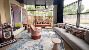 A modern living room with a sofa, armchairs, a coffee table, and a jukebox. Large windows provide a view of the outdoors. - ibis styles Paris Gennevilliers (Gennevilliers)