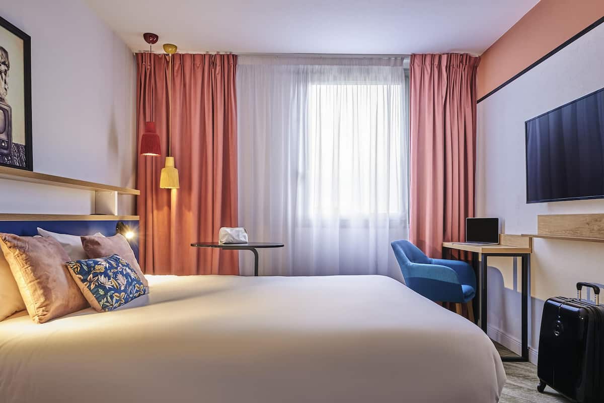 Une chambre d’hôtel avec un grand lit, un bureau, une chaise, une télévision et une valise.