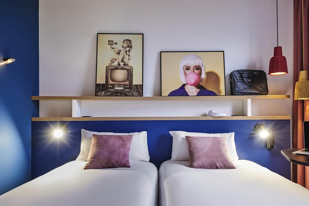 Une chambre d’hôtel avec deux lits, un téléviseur posé sur une étagère, un lampadaire rouge suspendu et un sac à main noir sur l’étagère.