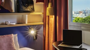 A hotel room with a bed, a laptop on a small table, a smartphone, a handbag, and a city view through the window. - ibis styles Paris Gennevilliers (Gennevilliers)