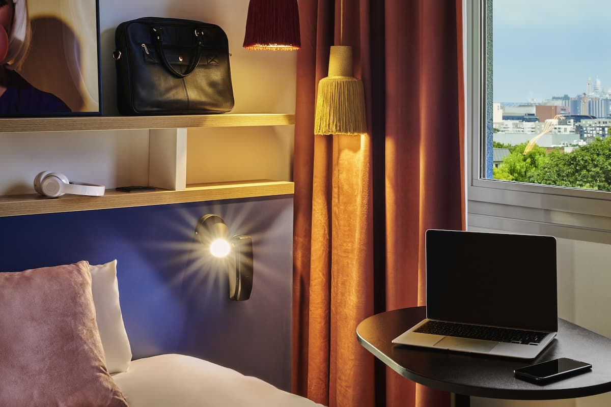 Une chambre d’hôtel avec un lit, un ordinateur portable posé sur une petite table, un téléphone intelligent, un sac à main et une vue sur la ville par la fenêtre.