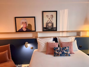 A hotel room with a bed, pillows, a nightstand, and a wall-mounted light. - ibis styles Paris Gennevilliers (Gennevilliers)