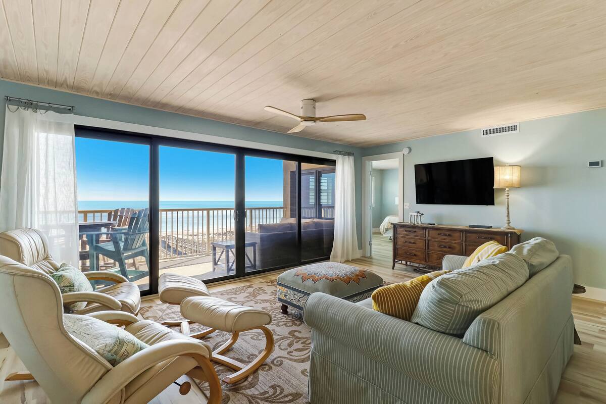 A spacious living room with a large sliding glass door leading to a balcony with a view of the ocean. There is a ceiling fan, a flat-screen TV, and a wooden cabinet.