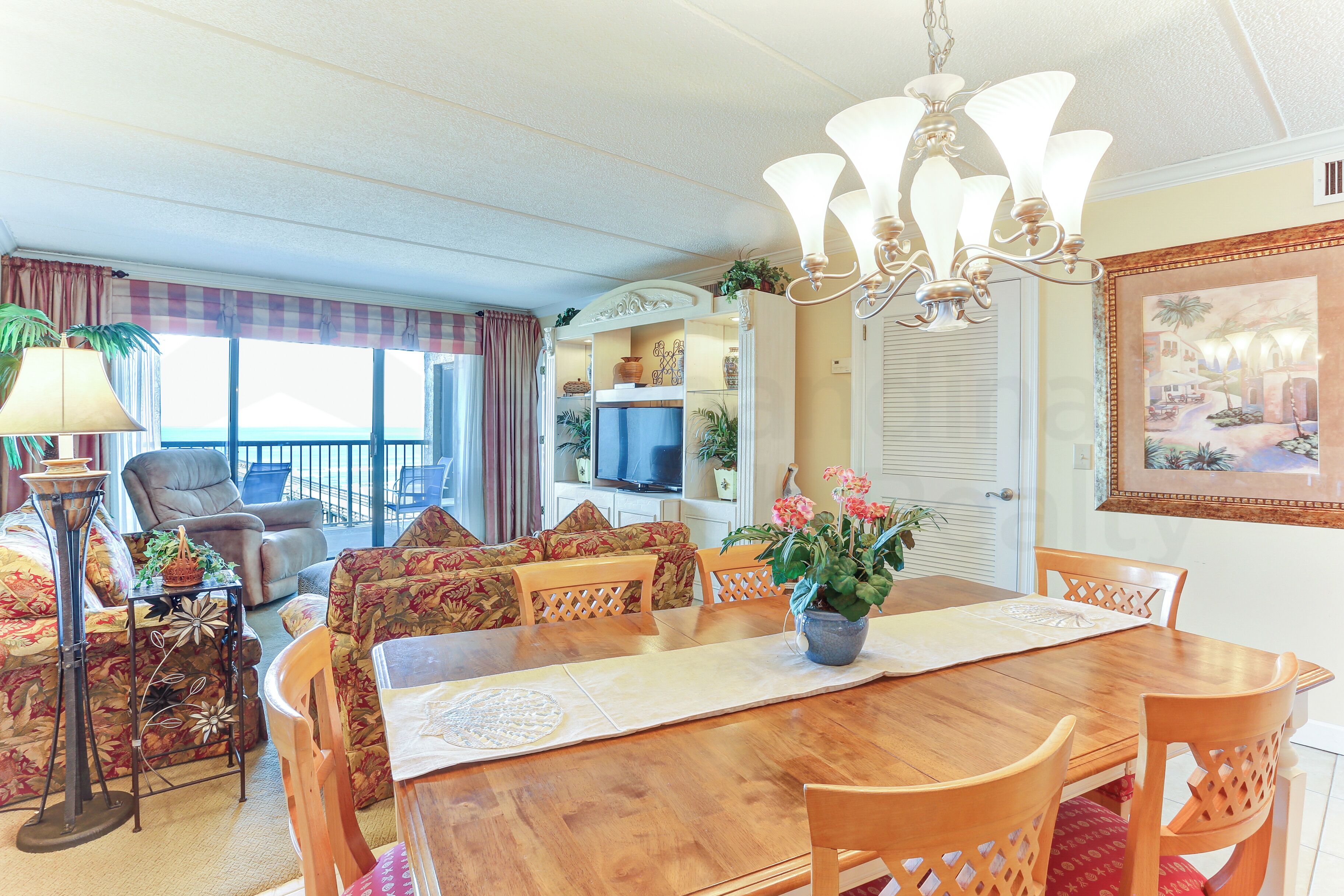 A dining area with a wooden table, chairs, and a chandelier. There is a television in the background and a balcony with a view.