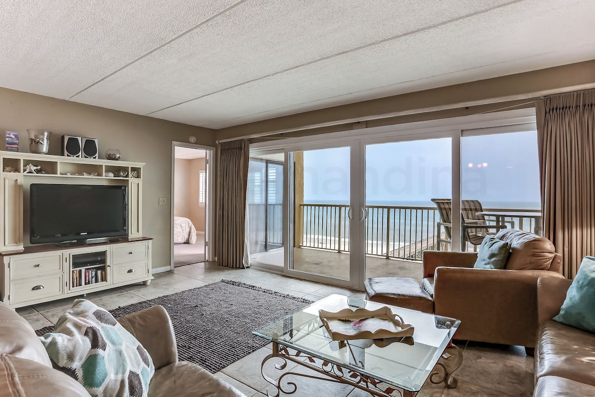 A living room with a flat-screen TV, a glass coffee table, and a sofa. There is a balcony with a view of the sea.