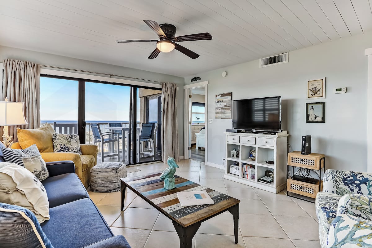 A living room with a sofa, coffee table, and TV stand. There is a balcony with a view.