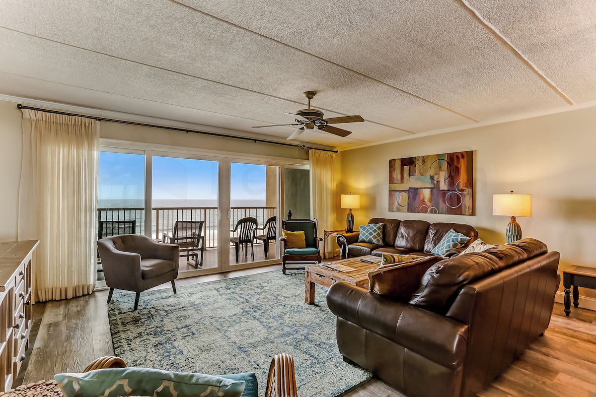 A spacious living room with a large leather sofa, a wooden coffee table, and a ceiling fan. There is a view of the outdoors through a sliding glass door.