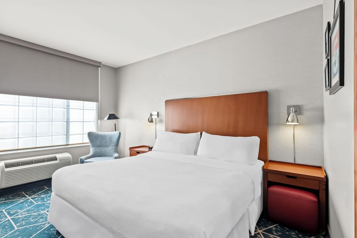 Ein Hotelzimmer mit einem großen Bett, einem Nachttisch mit Lampe, einem Sessel und einem roten Koffer.
