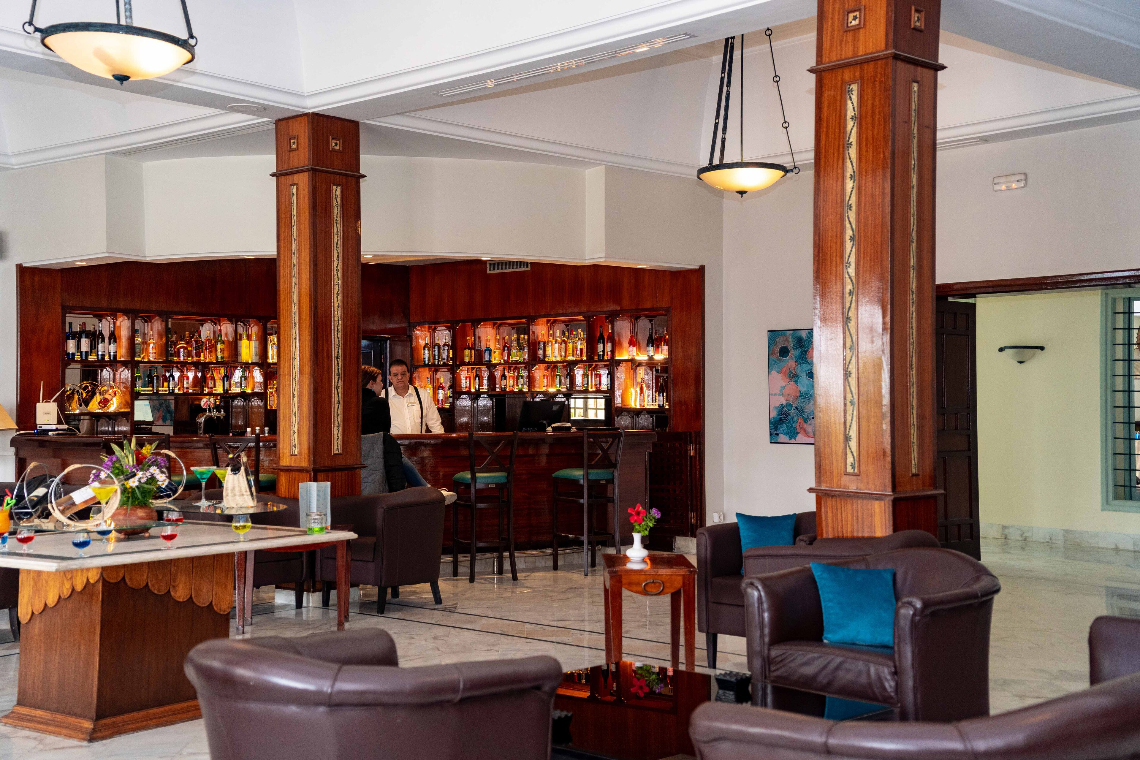 A bar area with a wooden bar counter, multiple seating options including leather chairs and a small table, and a variety of bottles and glasses on shelves.