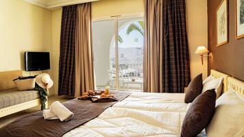 A hotel room with a bed, a TV, a balcony with curtains, and a view of palm trees.