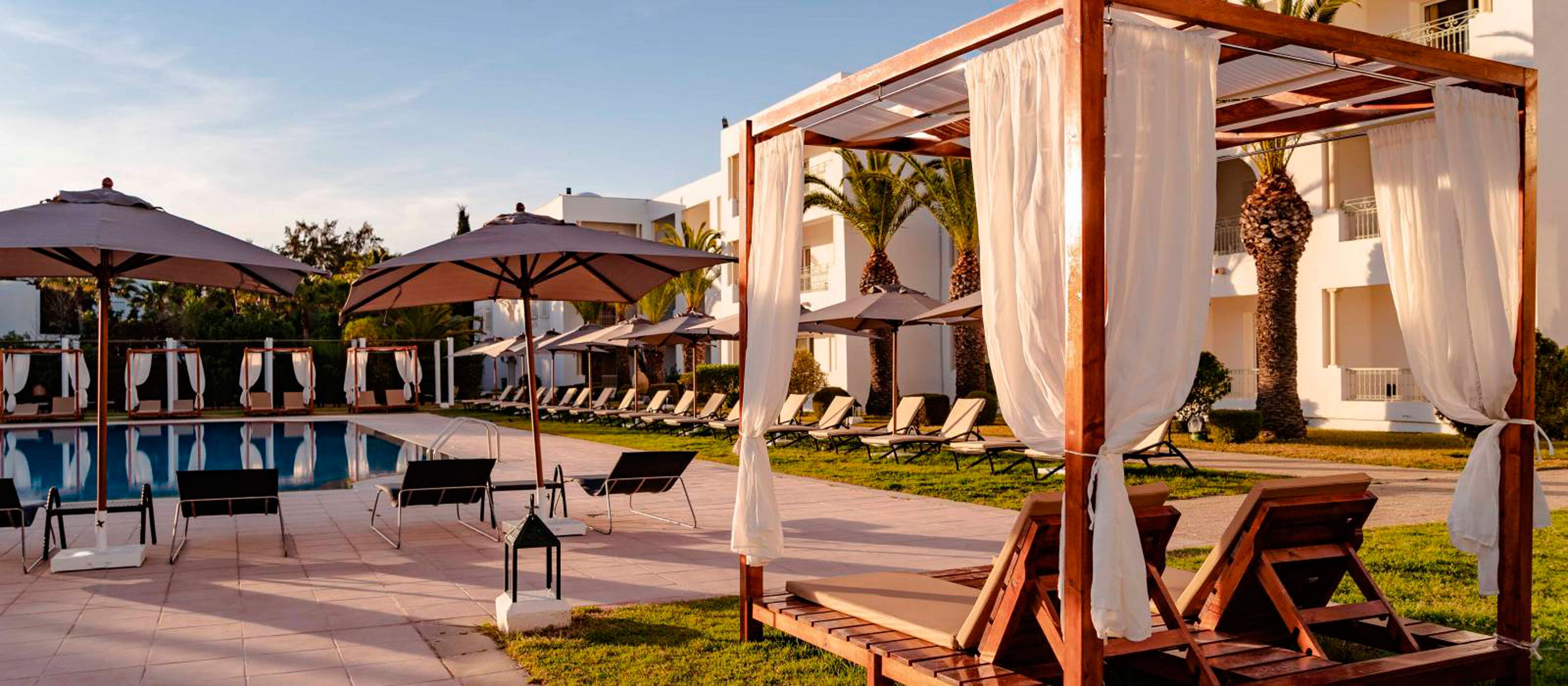A poolside area with sun loungers, umbrellas, and a pergola with white curtains.