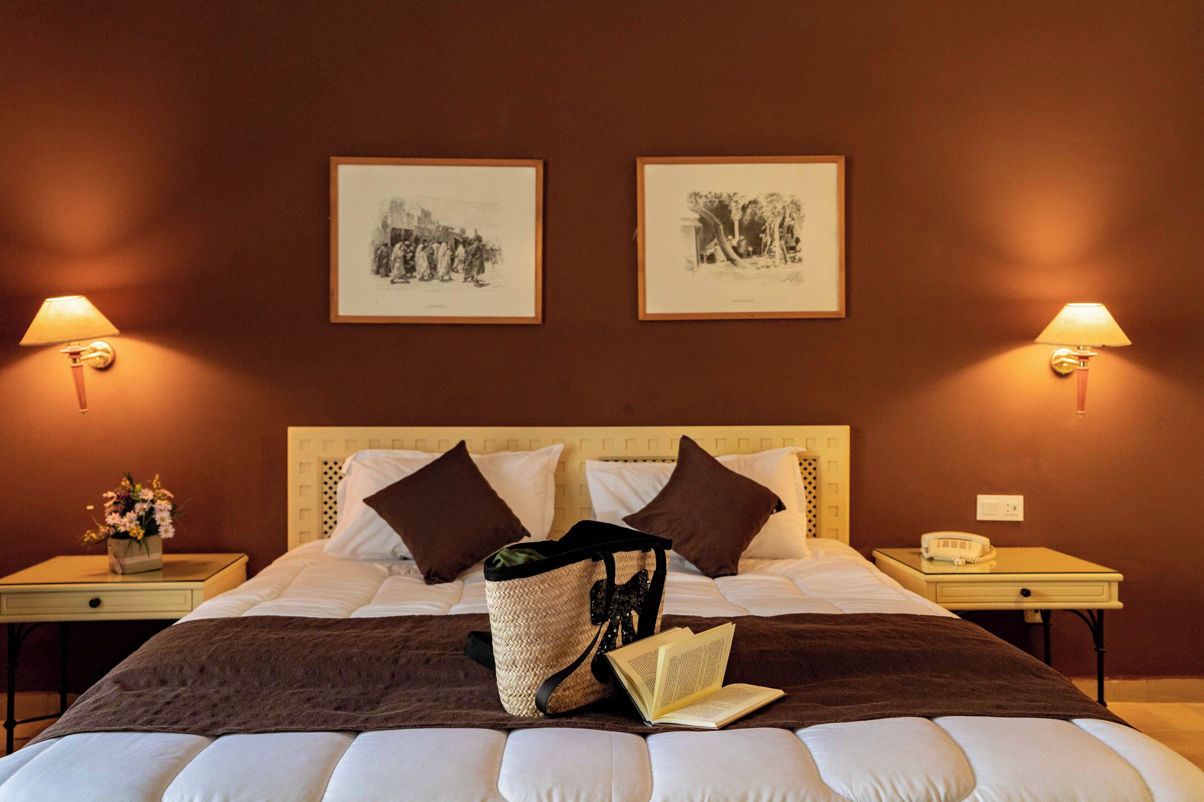 Une chambre d’hôtel avec un lit, deux tables de chevet, une lampe, un tableau encadré et un sac contenant un livre.