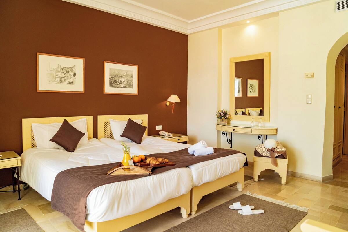 Quarto de hotel com uma cama grande, mesa de cabeceira com abajur, penteadeira com espelho e obras de arte nas paredes.