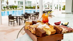 Ein Buffet mit verschiedenen Brotsorten und Gebäck, ein Getränk mit Trinkhalm und eine Obstschale auf einem Tisch in einem sonnigen Außenbereich mit Stühlen und einem Pool im Hintergrund.