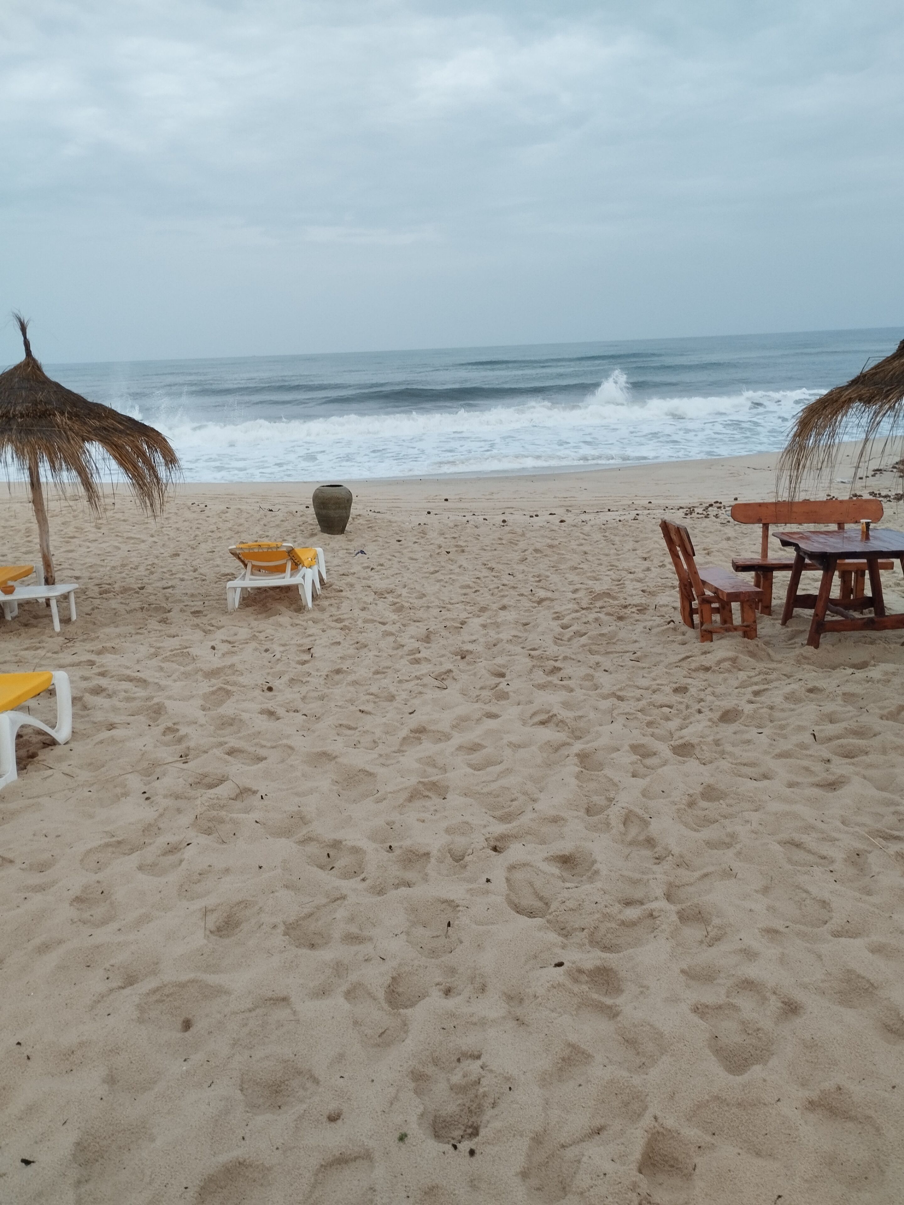 A beach with white and yellow lounge chairs, a wooden table, and a thatched umbrella.