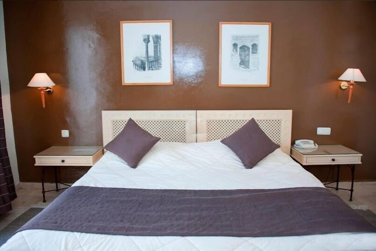 Uma cama de casal com duas almofadas, uma mesa de cabeceira com abajur e duas imagens emolduradas na parede.