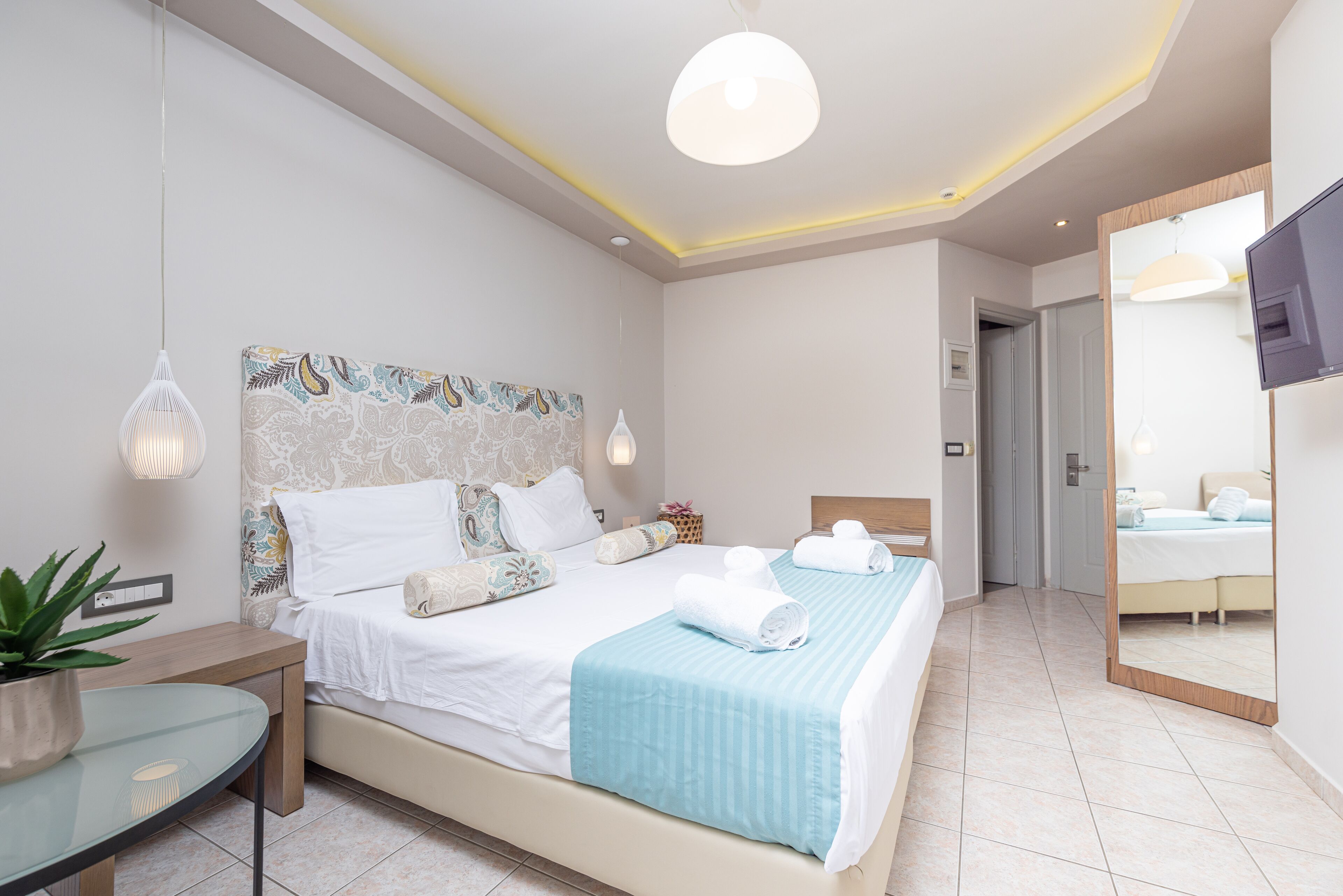 A modern hotel room with a large bed, bedside tables, a potted plant, and a TV.