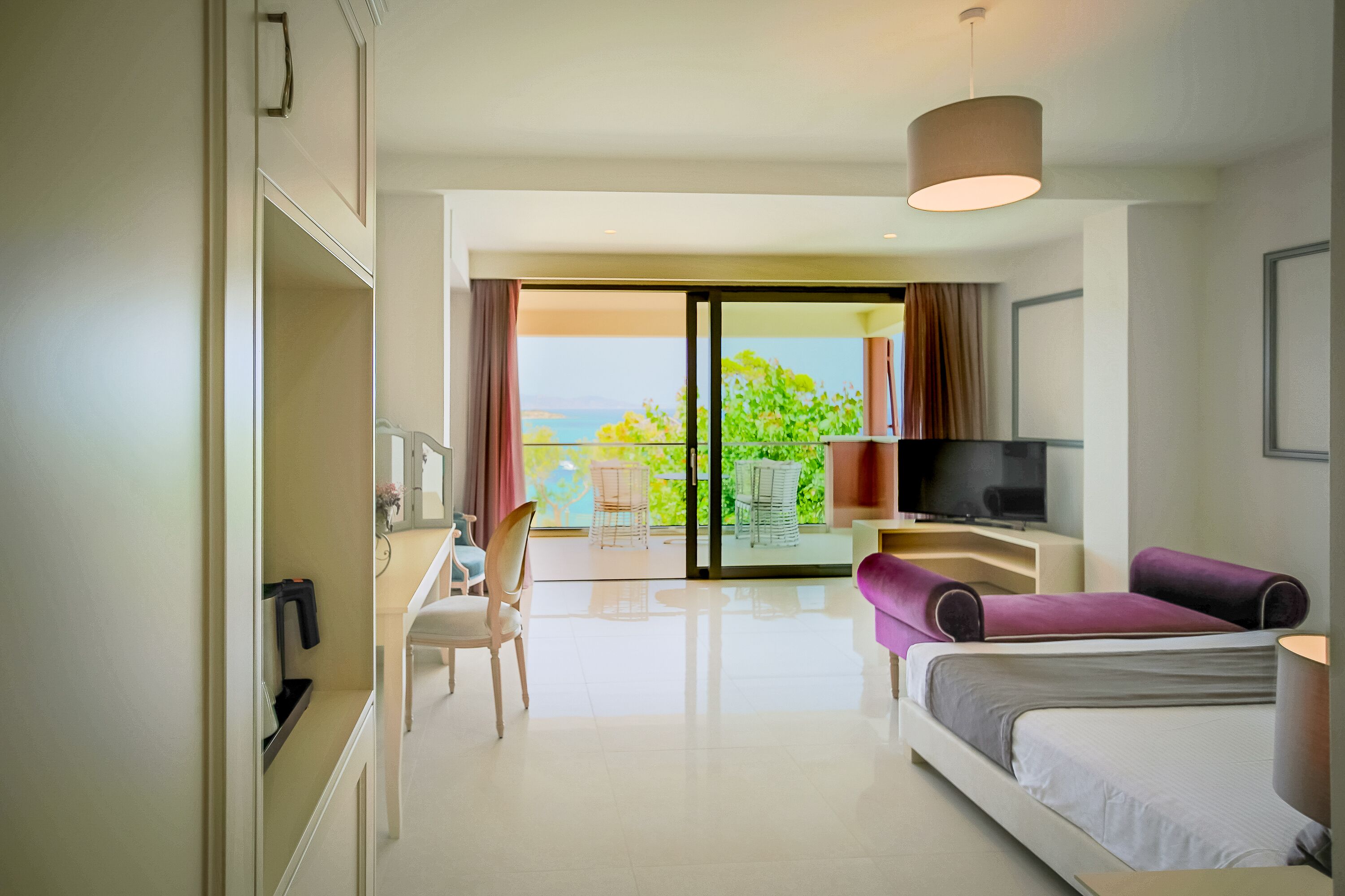 A modern hotel room with a bed, a desk, a TV, and a balcony with a view of the sea.
