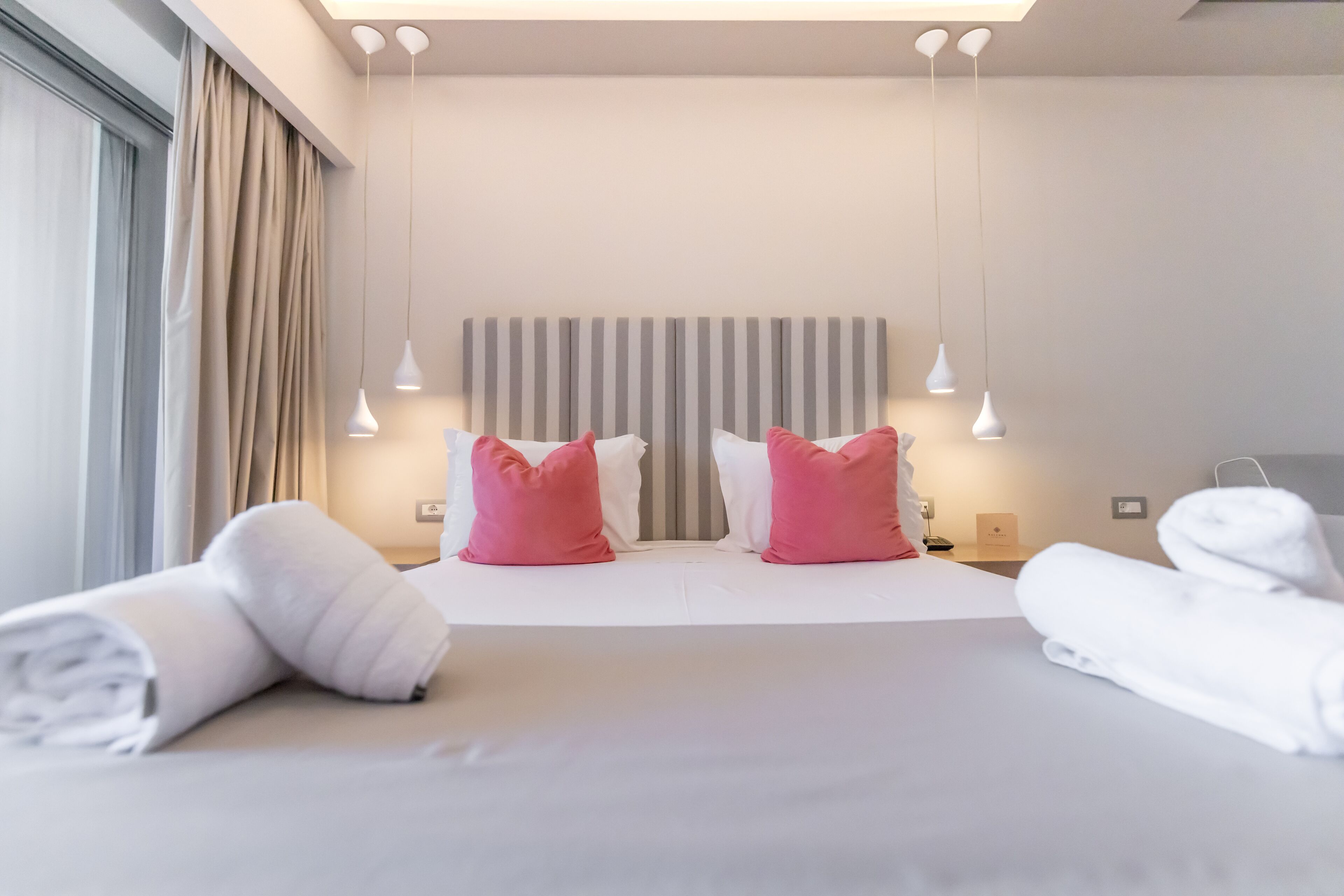A modern hotel room with a large bed, two pink pillows, white towels, and a striped headboard.