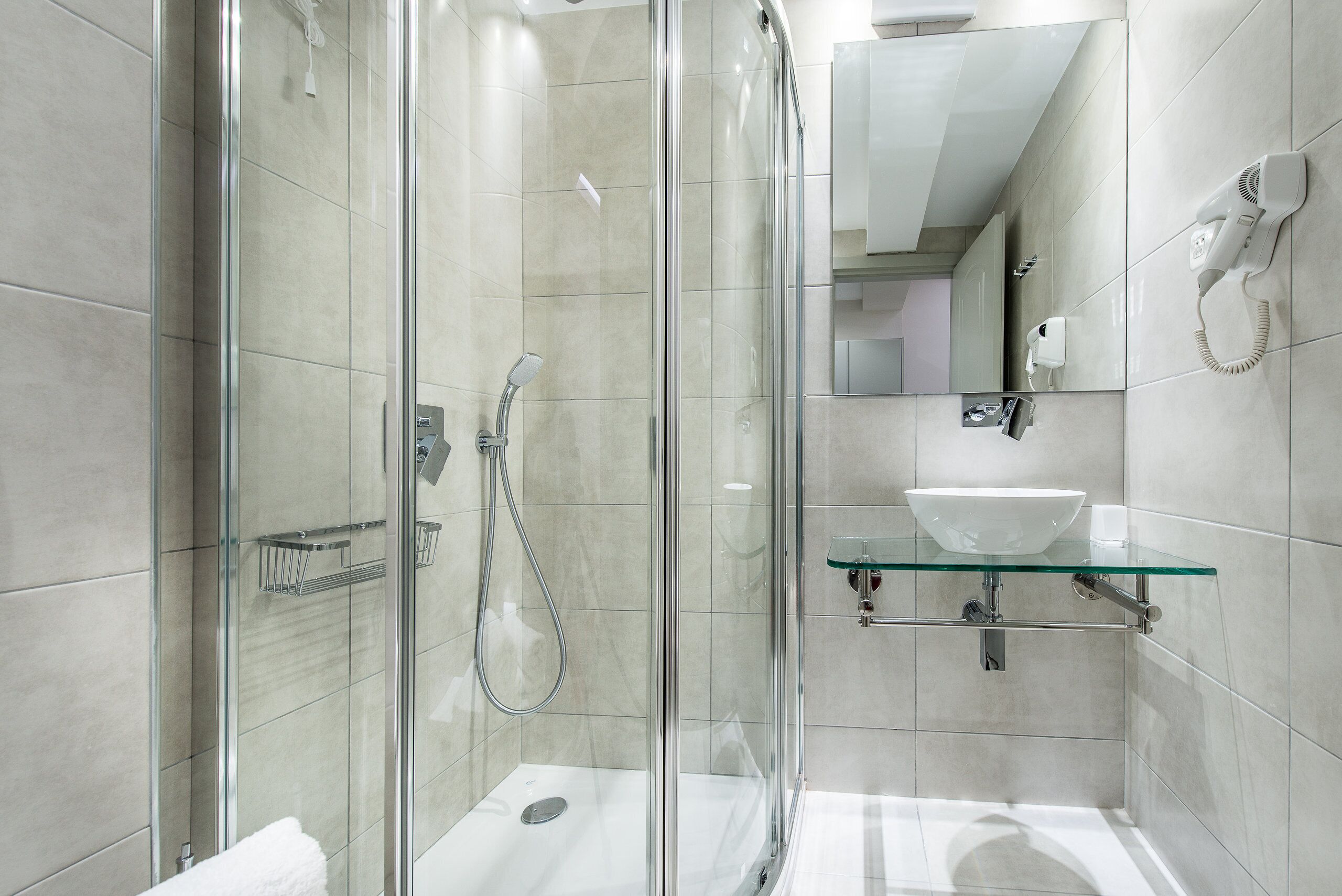Une salle de bain moderne dotée d’une douche à parois de verre, d’un lavabo en verre et d’un sèche-cheveux portatif fixé au mur.