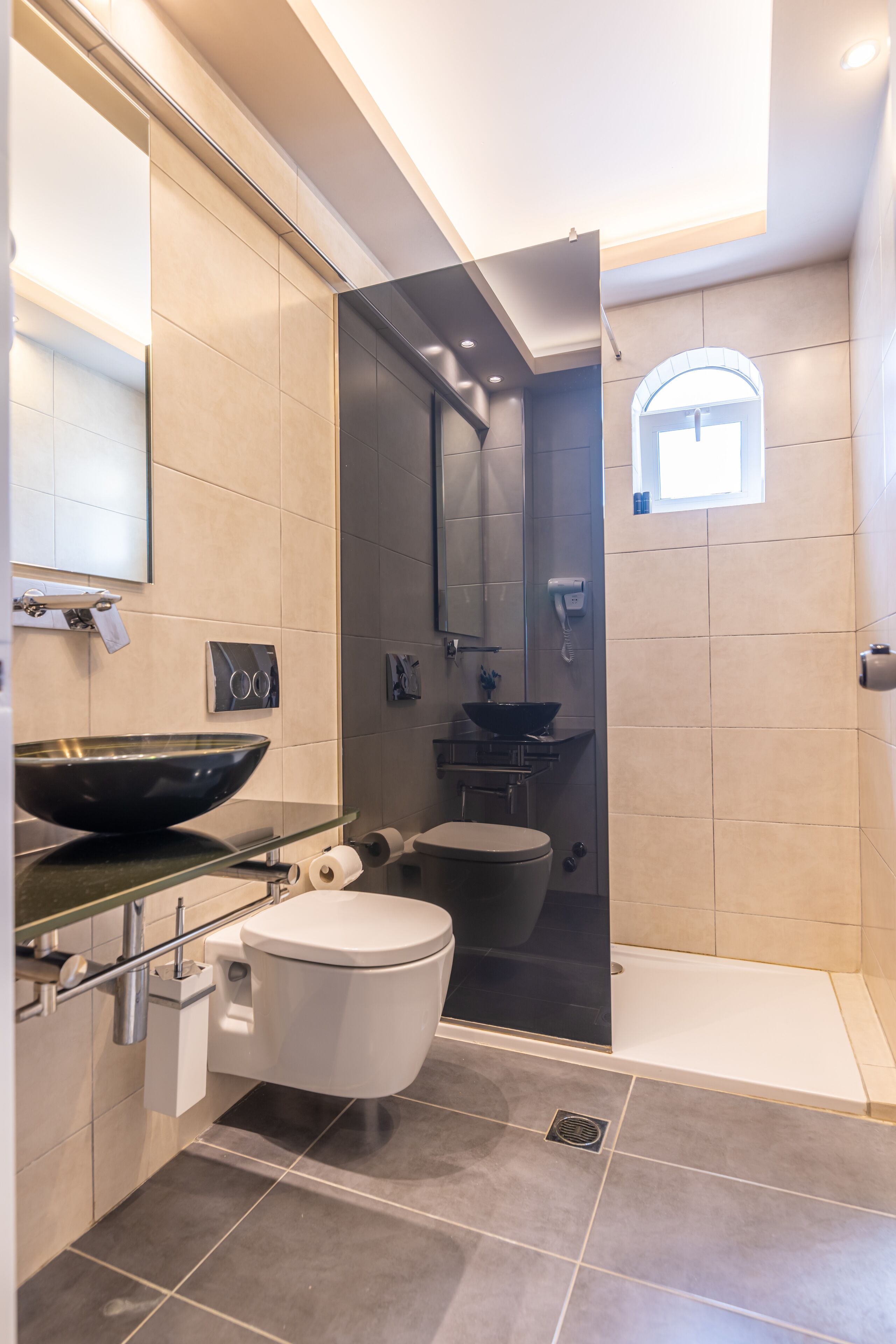 A modern bathroom with a glass shower enclosure, a white toilet, a black basin, and a small window above the sink.