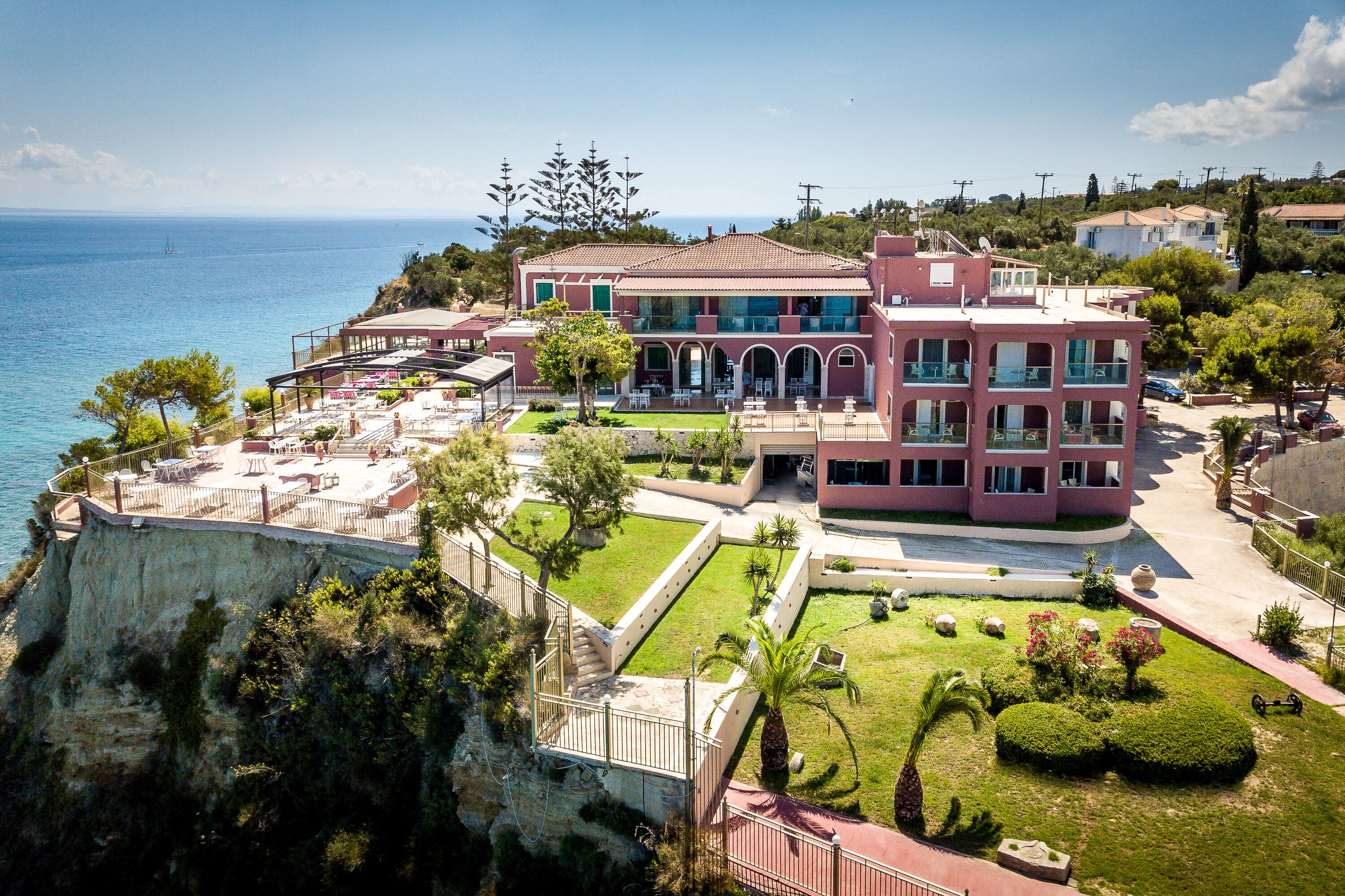 Un edificio grande de varios pisos con un balcón que da al mar, rodeado de jardines cuidados y un acantilado.