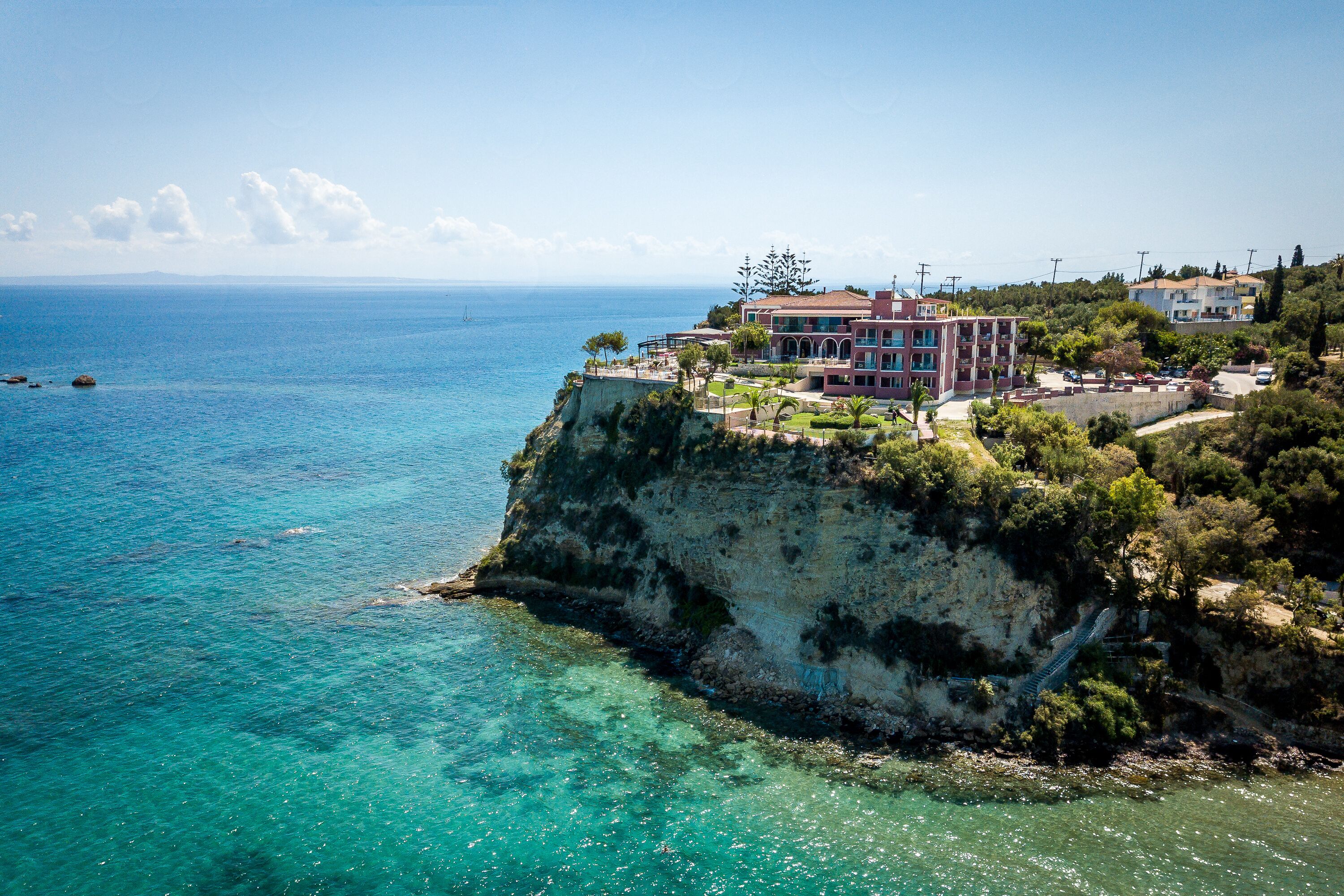 A coastal building with a balcony overlooking the sea, situated on a cliff with clear waters.