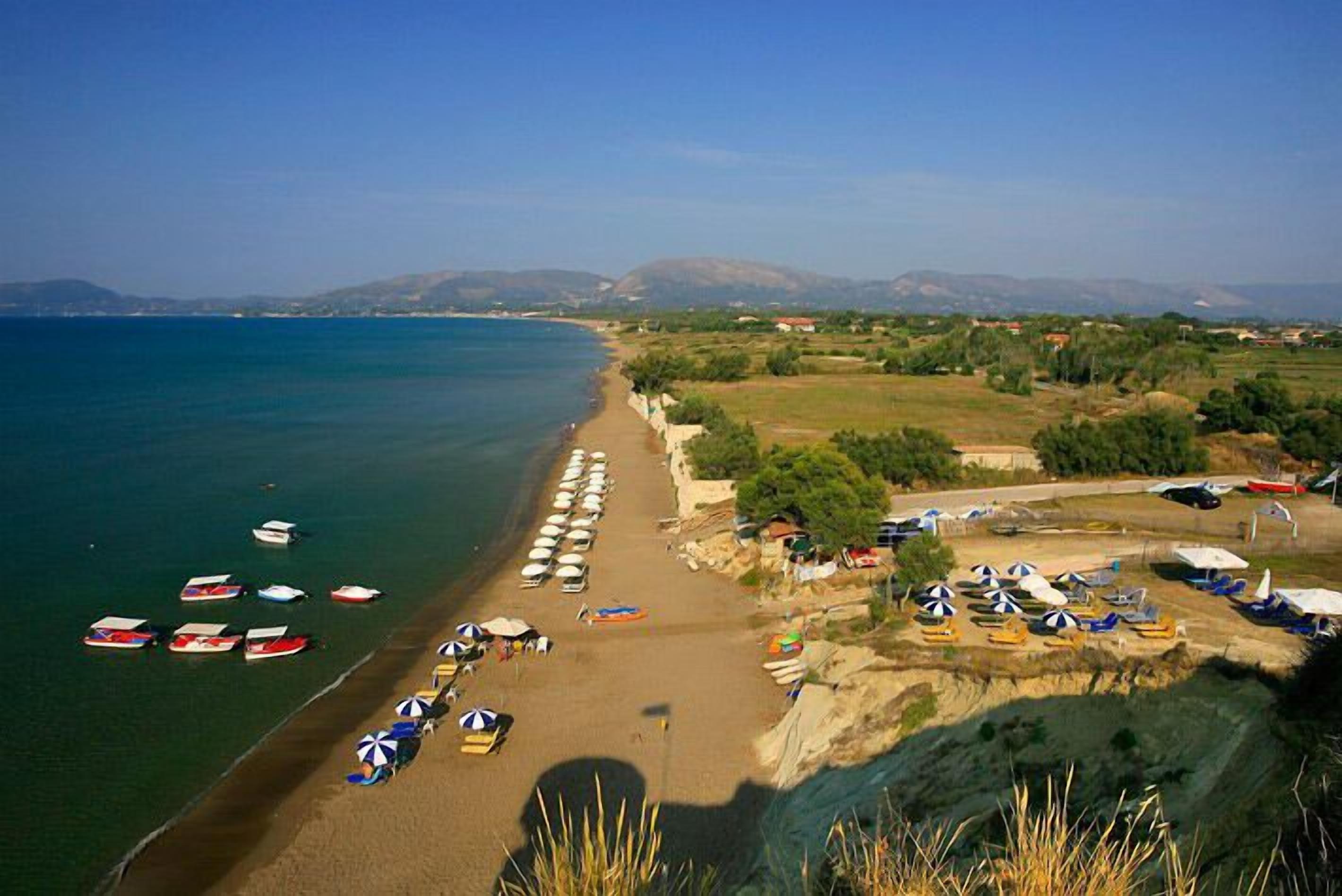 A coastal beach with boats, umbrellas, and a clear view of the sea and distant hills.
