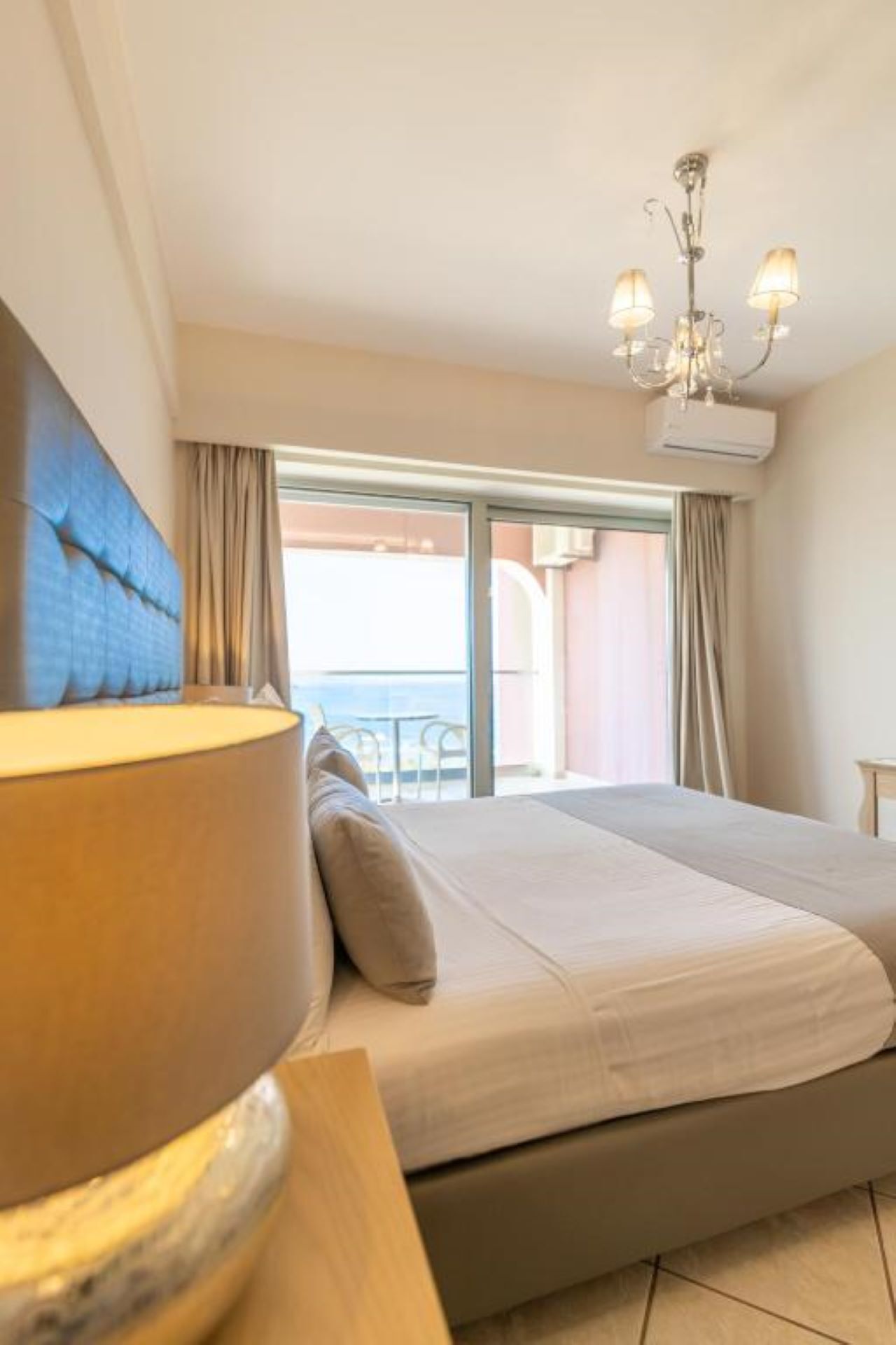 A hotel room with a bed, a lamp, a view of the sea, and a balcony.