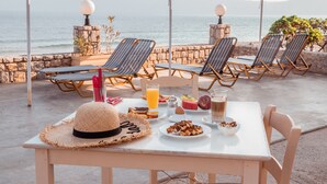 A table with breakfast items, a straw hat, and lounge chairs by a seaside view. - Galini Beach Hotel (Kissamos)