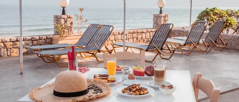 A table with breakfast items, a straw hat, and lounge chairs by a seaside view.