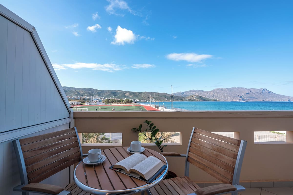 A balcony with a table, chairs, and a view of the sea and mountains.