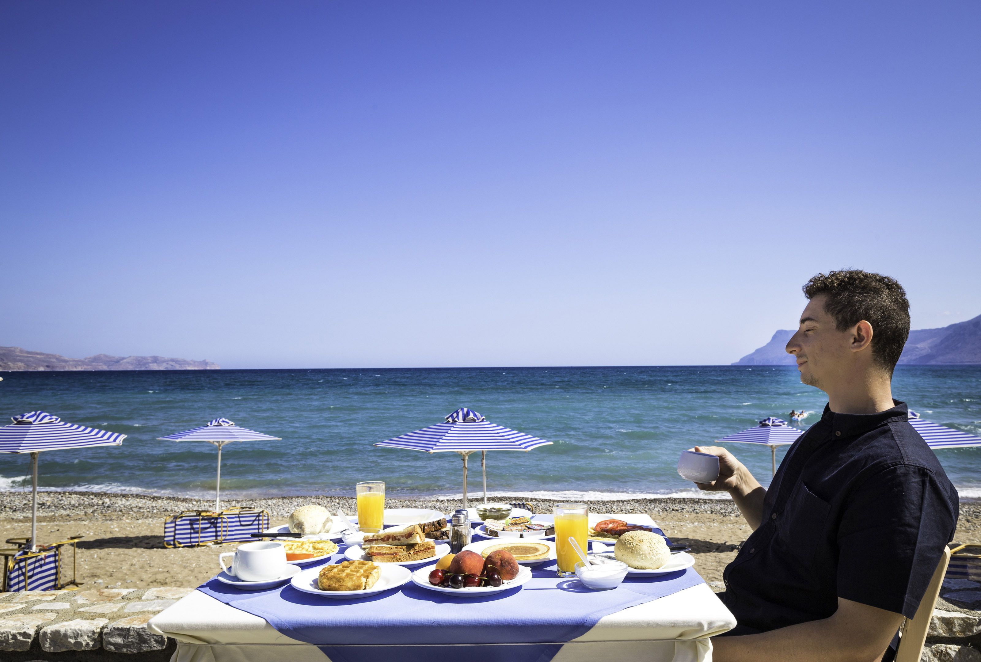 A table set with breakfast items on a beach with umbrellas and the sea in the background.