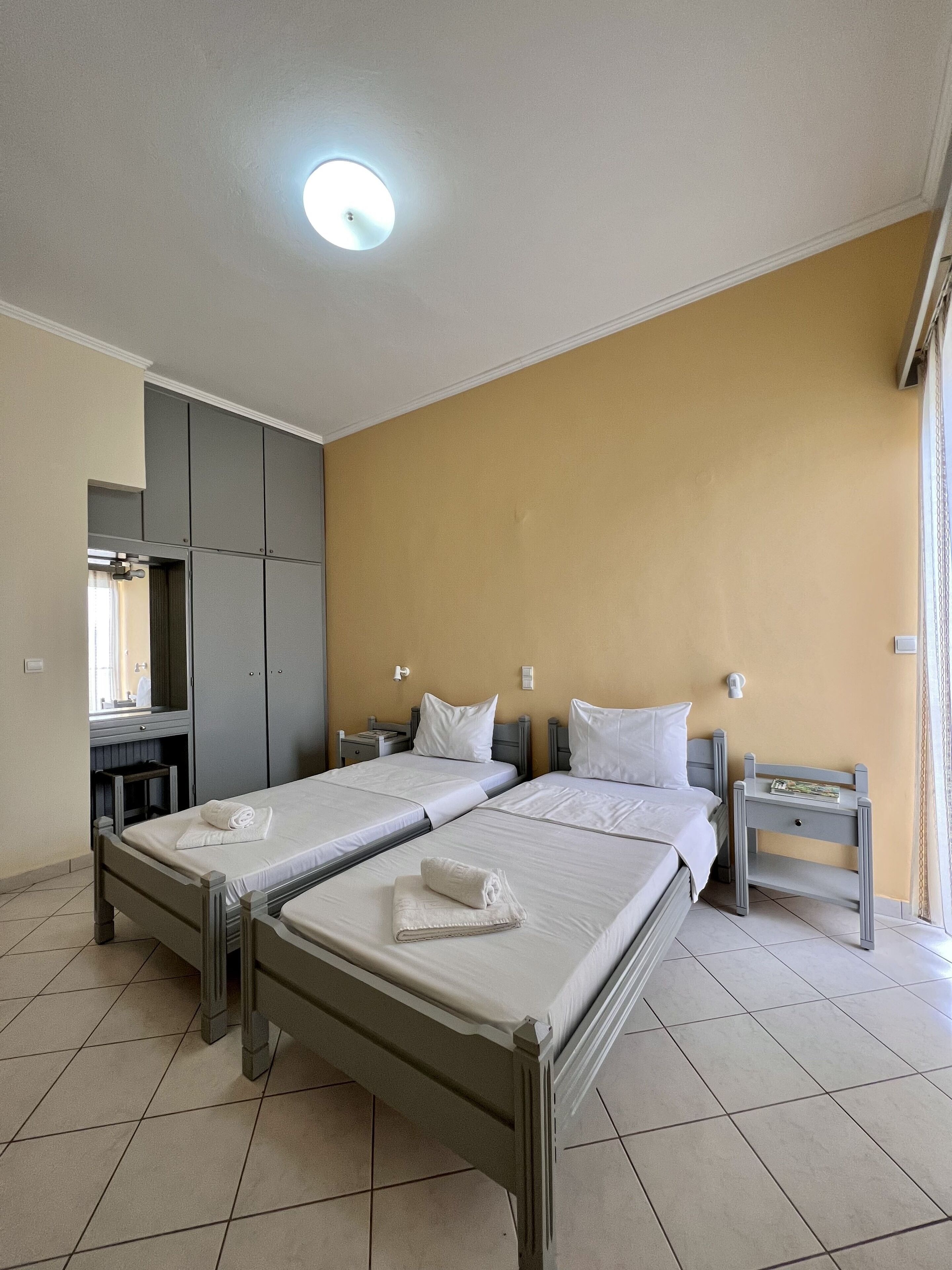 A hotel room with two beds, a wardrobe, a mirror, and a small table with a lamp.