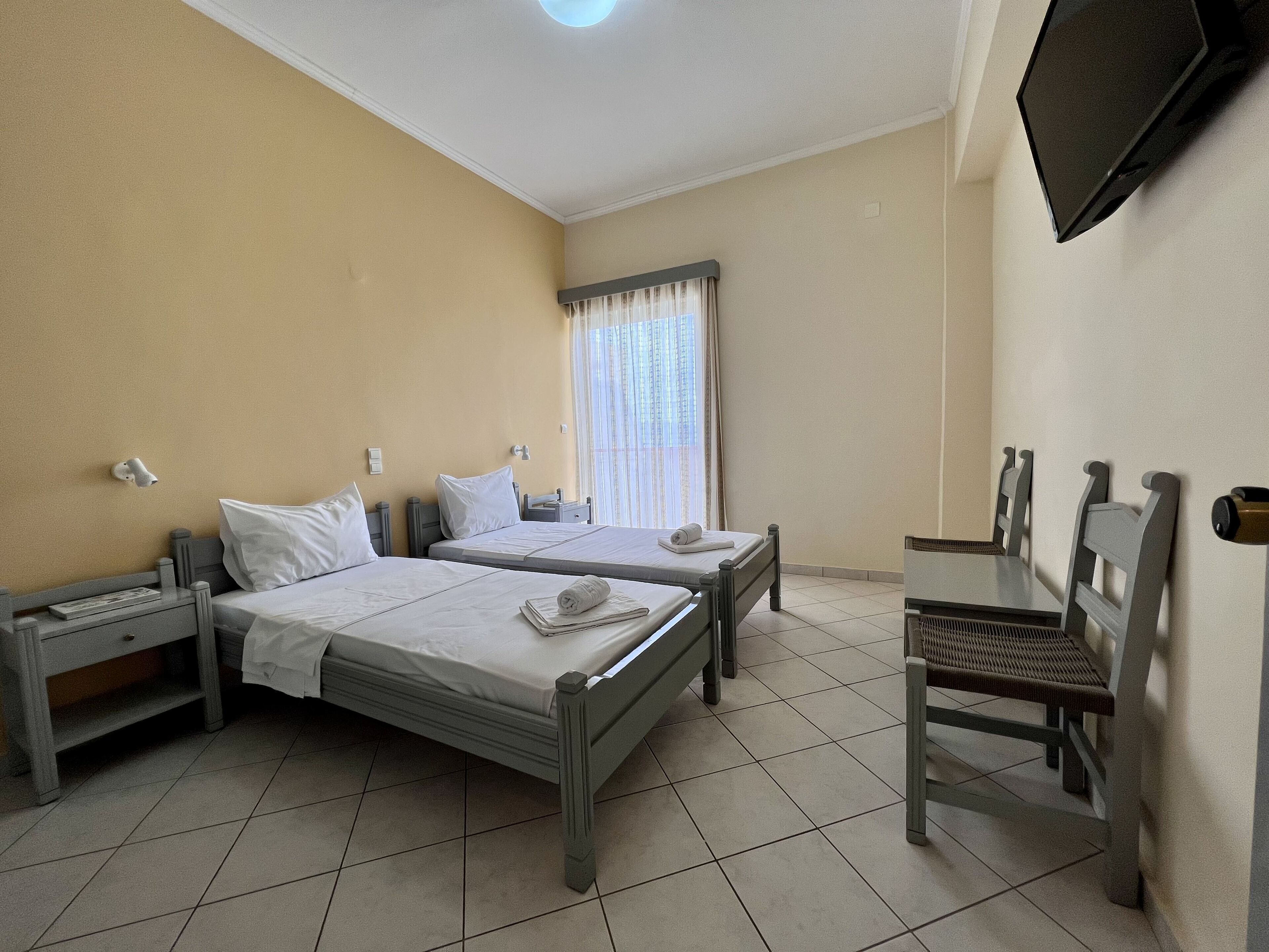 Habitación de hotel con dos camas individuales, un escritorio, una silla, un televisor y una ventana con cortinas.