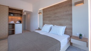 A modern hotel room with a large bed, a wooden headboard, a bedside table with a plant, and a built-in wardrobe with a small refrigerator and a radio. - Galini Beach Hotel (Kissamos)