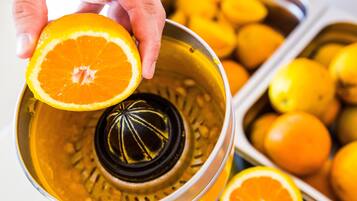 Una mano sostiene media naranja sobre un exprimidor, con naranjas al fondo.