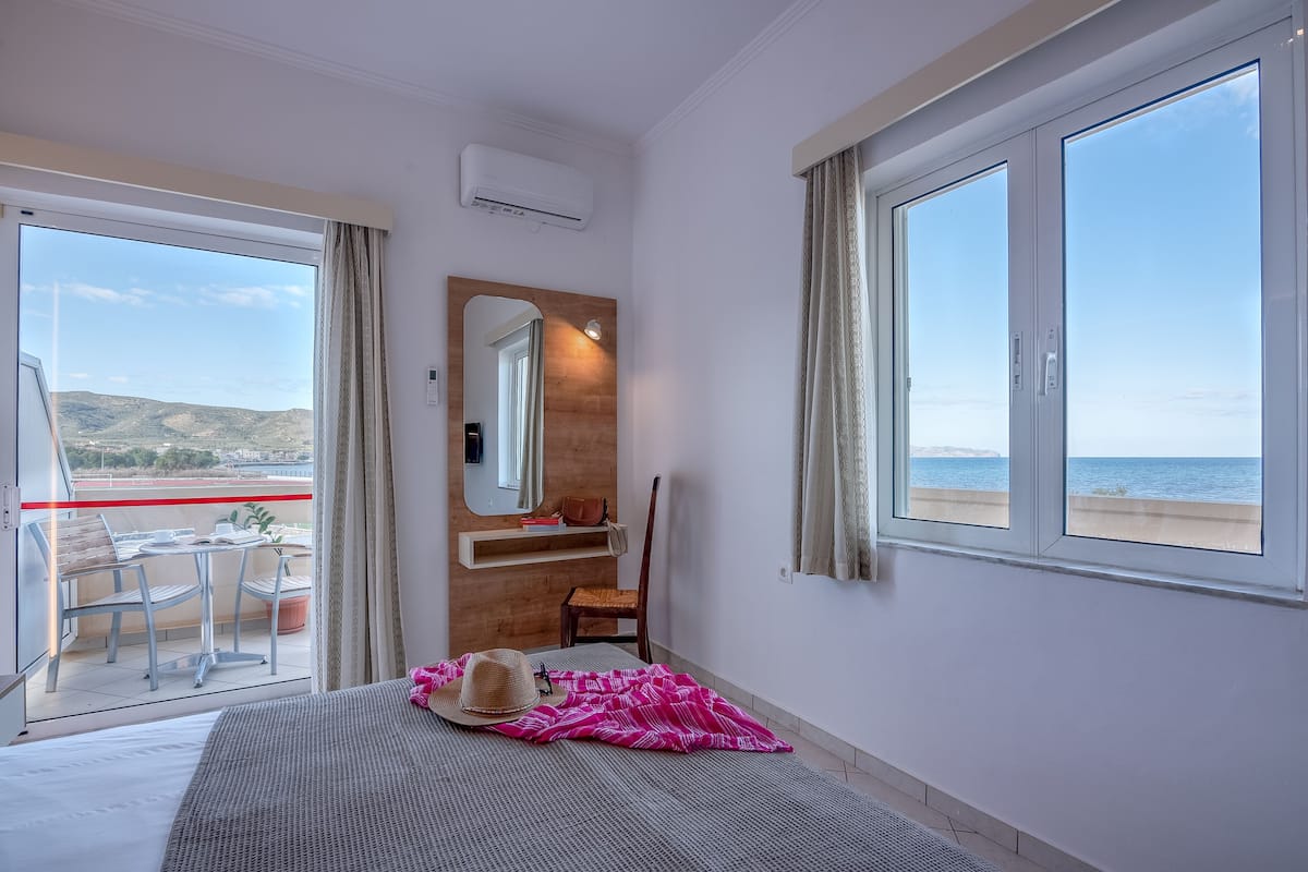 A hotel room with a bed, a chair, a small table, a mirror, and a view of the sea.