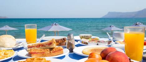 Una mesa servida con alimentos de desayuno, como waffles, tostadas, fruta y jugo, con vistas a un cuerpo de agua y sombrillas.