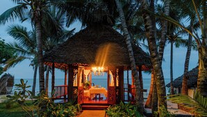Une construction à toit de chaume comprenant un espace repas, entourée de palmiers et offrant une vue sur la plage.