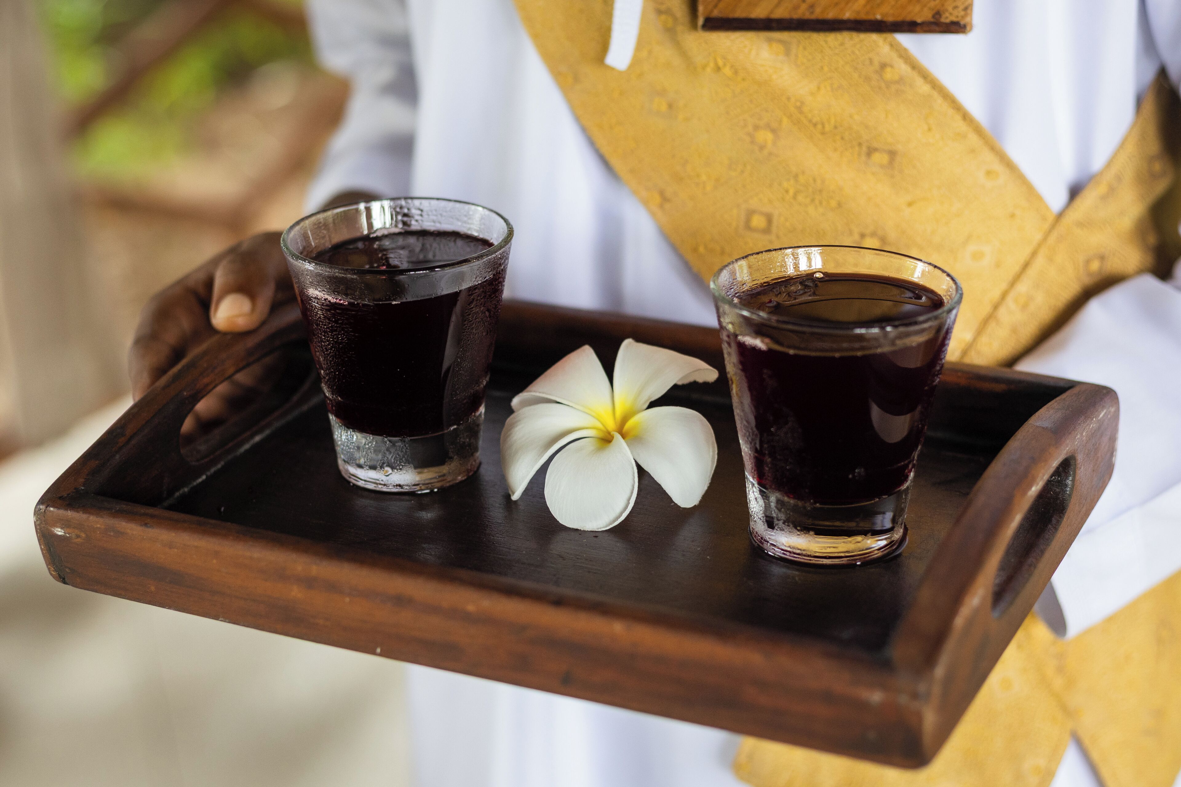 Una bandeja de madera con dos vasos de líquido oscuro, posiblemente una bebida, y una flor blanca.