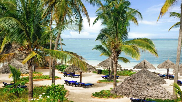 Un complexe hôtelier de bord de mer, avec des palmiers, des parasols en feuilles de palmier et des chaises longues.