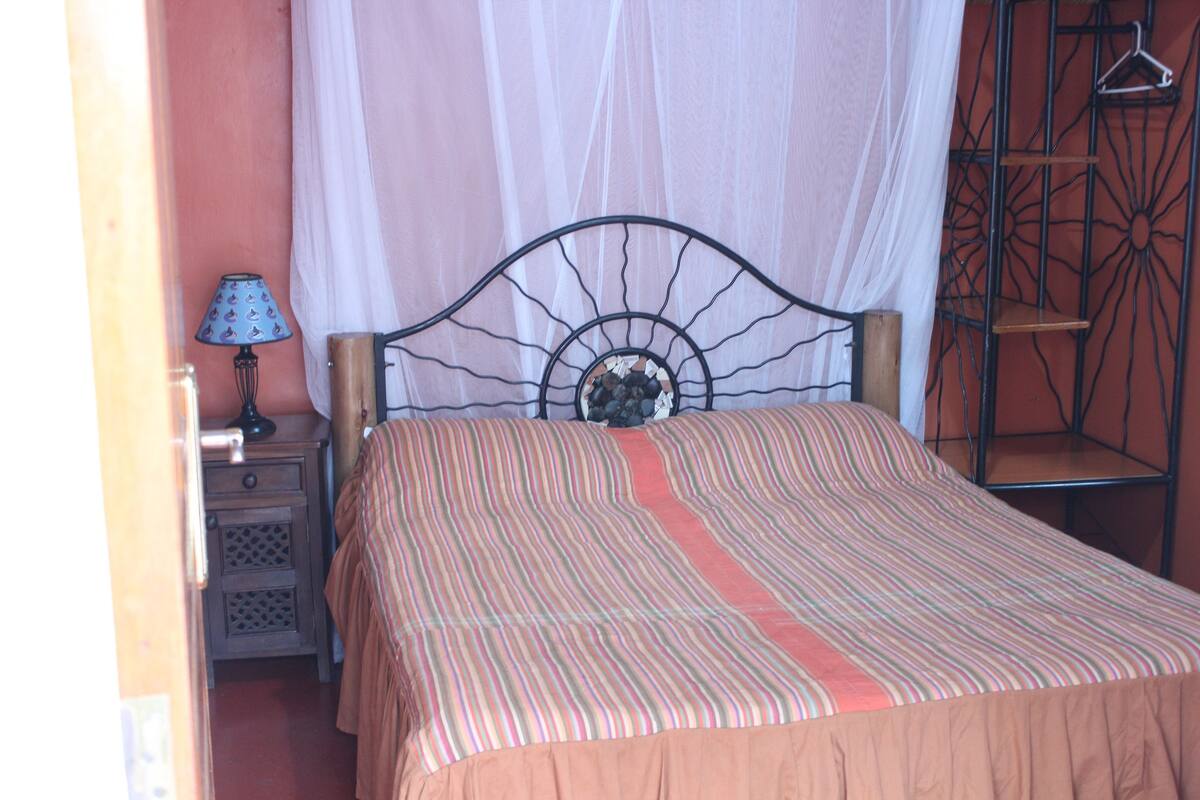 Una cama con cubrecama a rayas y dosel, una mesita de noche con lámpara, y una estantería metálica frente a una pared color melocotón.