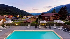 Une piscine entourée de bâtiments et de montagnes, avec des chaises longues et des parasols.