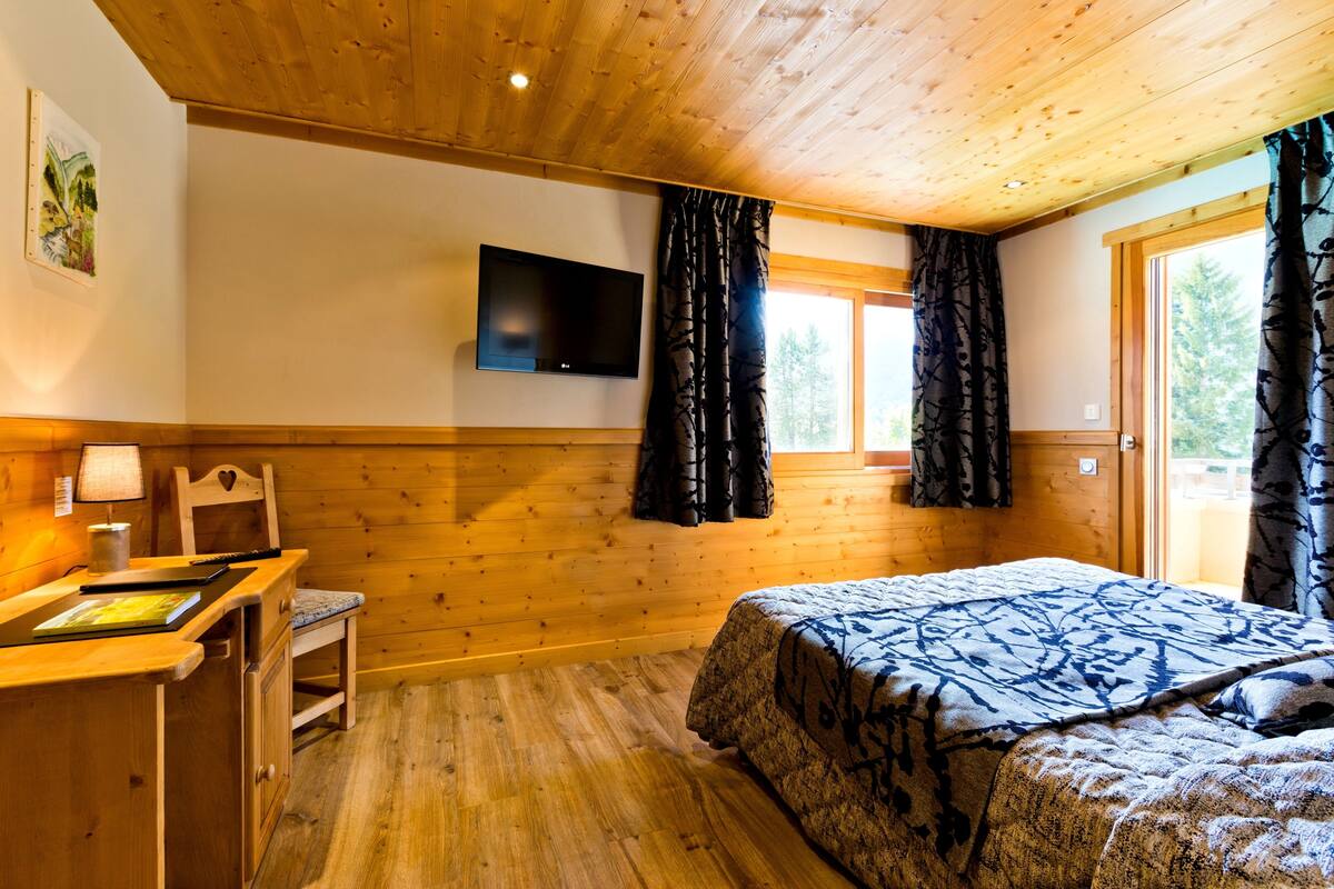 Une pièce de style cabane en bois, avec un lit, un bureau doté d’un ordinateur portable, un téléviseur fixé au mur et une fenêtre avec des rideaux.