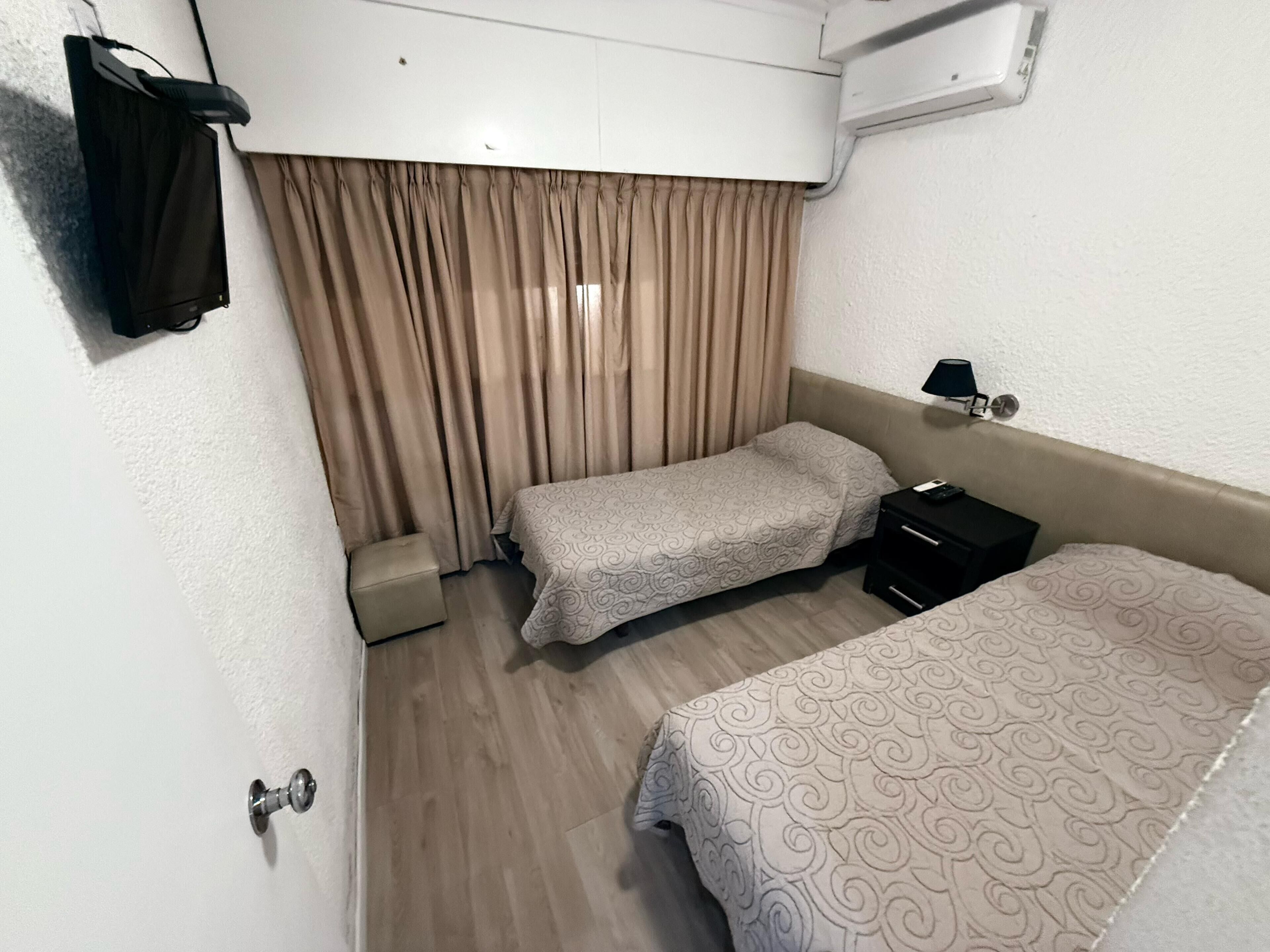A hotel room with two beds, a small bedside table with a lamp, a wall-mounted air conditioner, and a curtain.