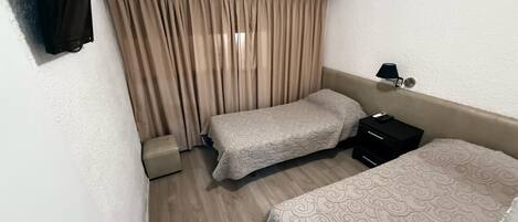 A hotel room with two beds, a small bedside table with a lamp, a wall-mounted air conditioner, and a curtain.